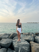 Model in the Caletta White 100% Cotton Pareo (short skirt style) posing on seaside rocks, ideal for beach resort wear.